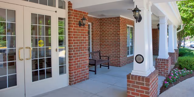 Covered brick entrance with white double glass doors, columns, a bench, lanterns, and landscaping.