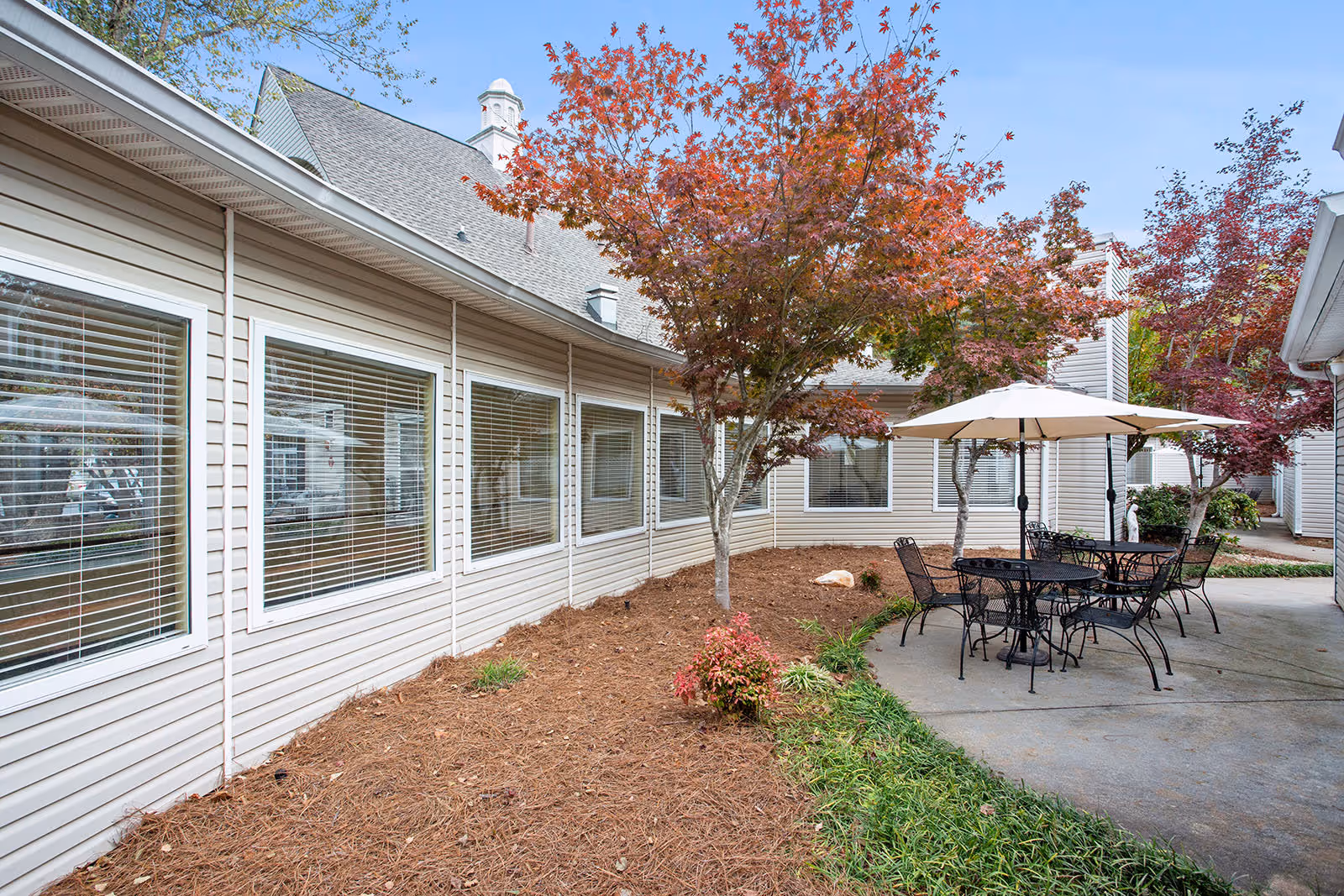 Outdoor patio area at Governors Glen featuring metal tables and chairs with large white umbrellas, surrounded by trees with red and orange autumn leaves, adjacent to a beige building with multiple windows.
