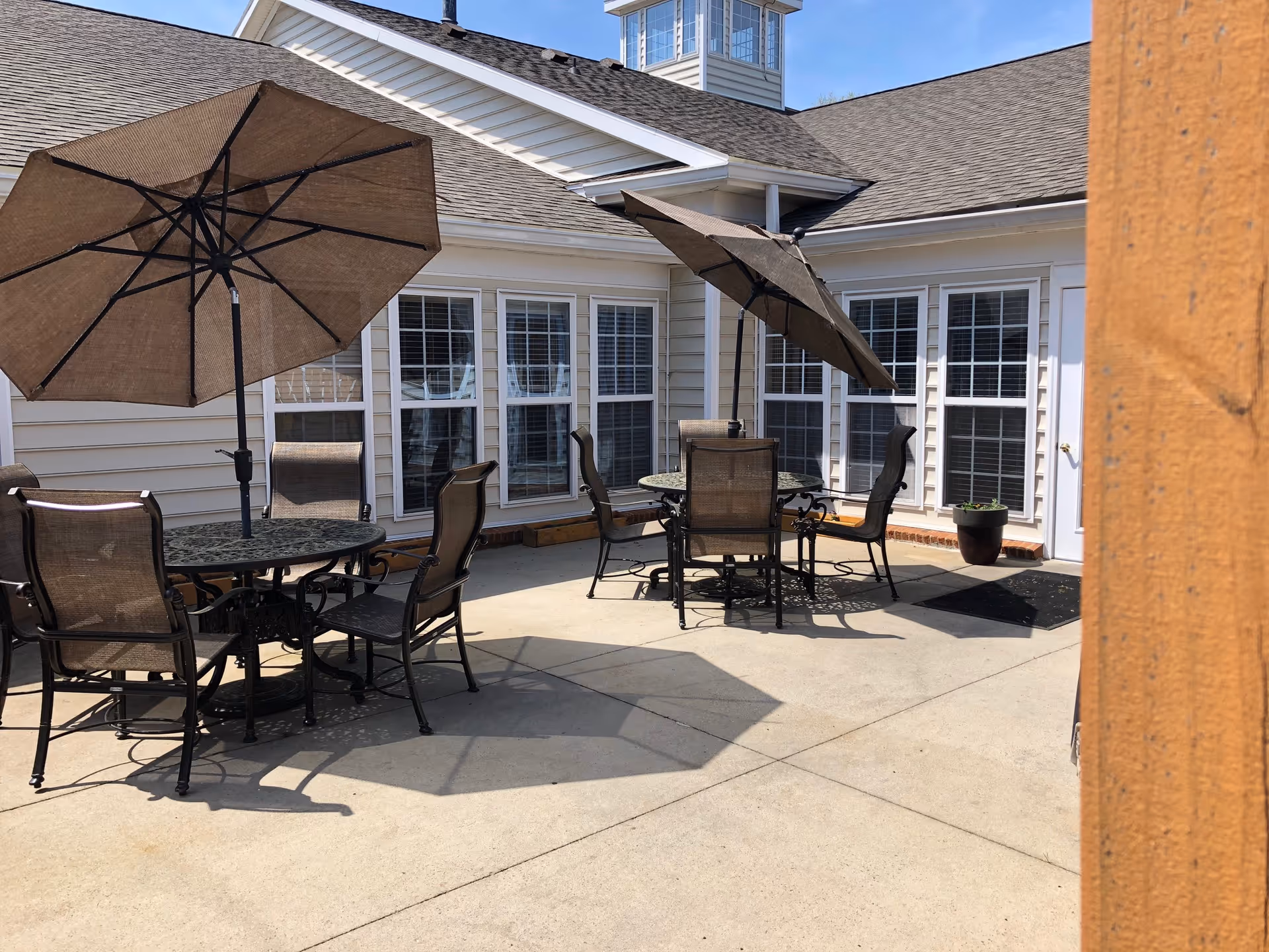 Sunny courtyard patio with round metal tables, chairs, and large umbrellas outside a beige assisted living building.