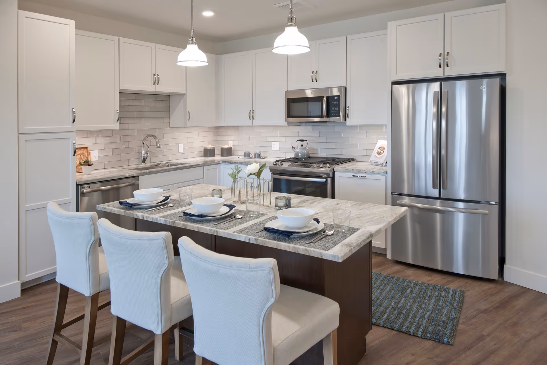 Modern kitchen with white cabinets, stainless steel appliances including a refrigerator, oven, and microwave. A kitchen island with a marble countertop is set with three place settings, each with a bowl, plate, napkin, and glass. Three upholstered chairs are positioned at the island. Two pendant lights hang above the island, and a small decorative plant and candles are on the countertop.