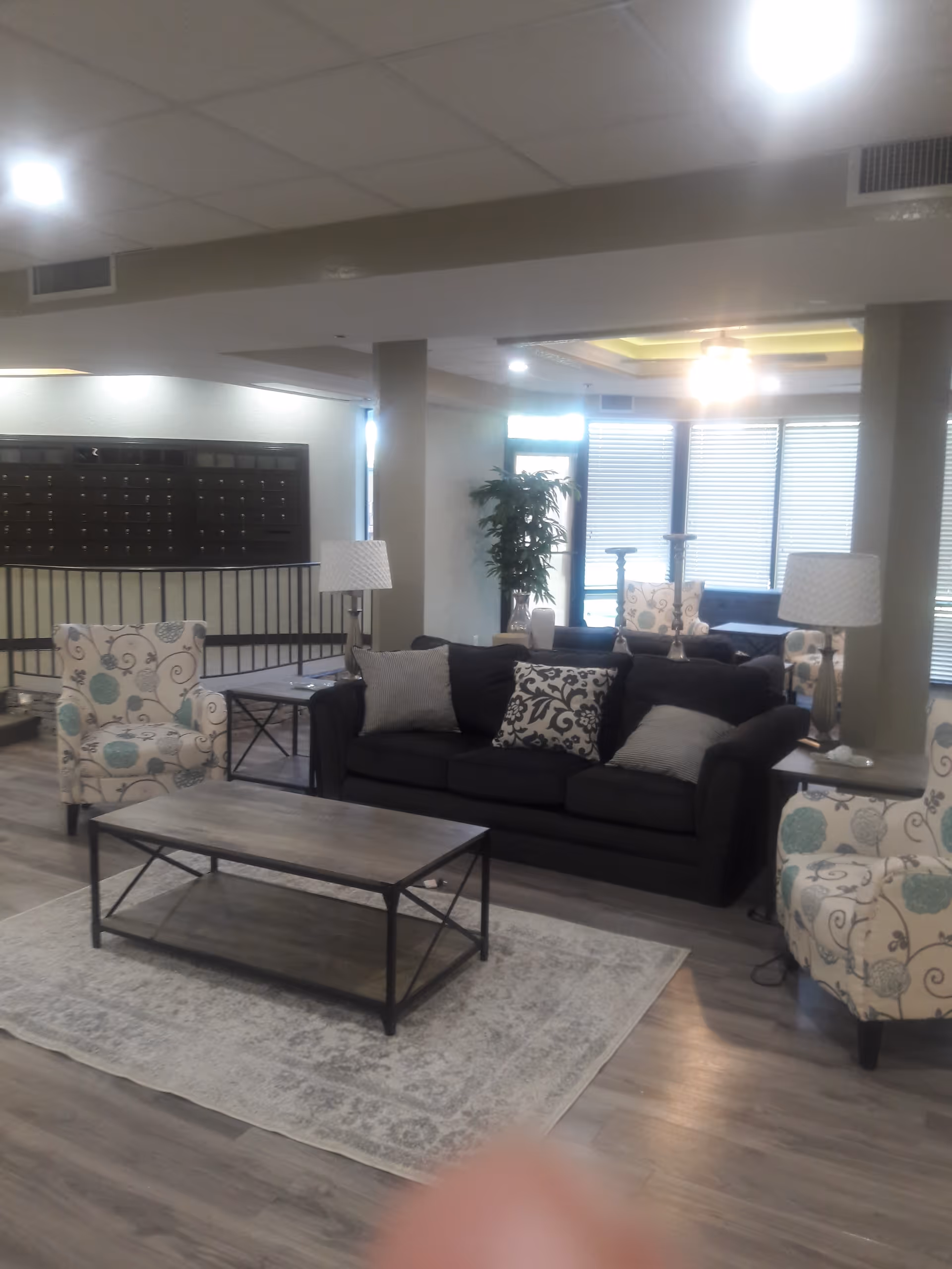 Furnished common living area with a dark sofa, patterned armchairs, coffee table, lamps, and mailboxes in the background.