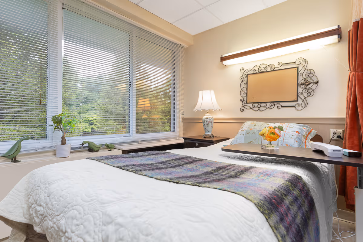 A cozy bedroom in a senior living facility with a neatly made bed covered in a white quilt and a colorful throw blanket. A bedside table holds a decorative lamp and a small vase with flowers. Large windows with blinds allow natural light to fill the room, and a decorative framed corkboard hangs on the wall above the bed.