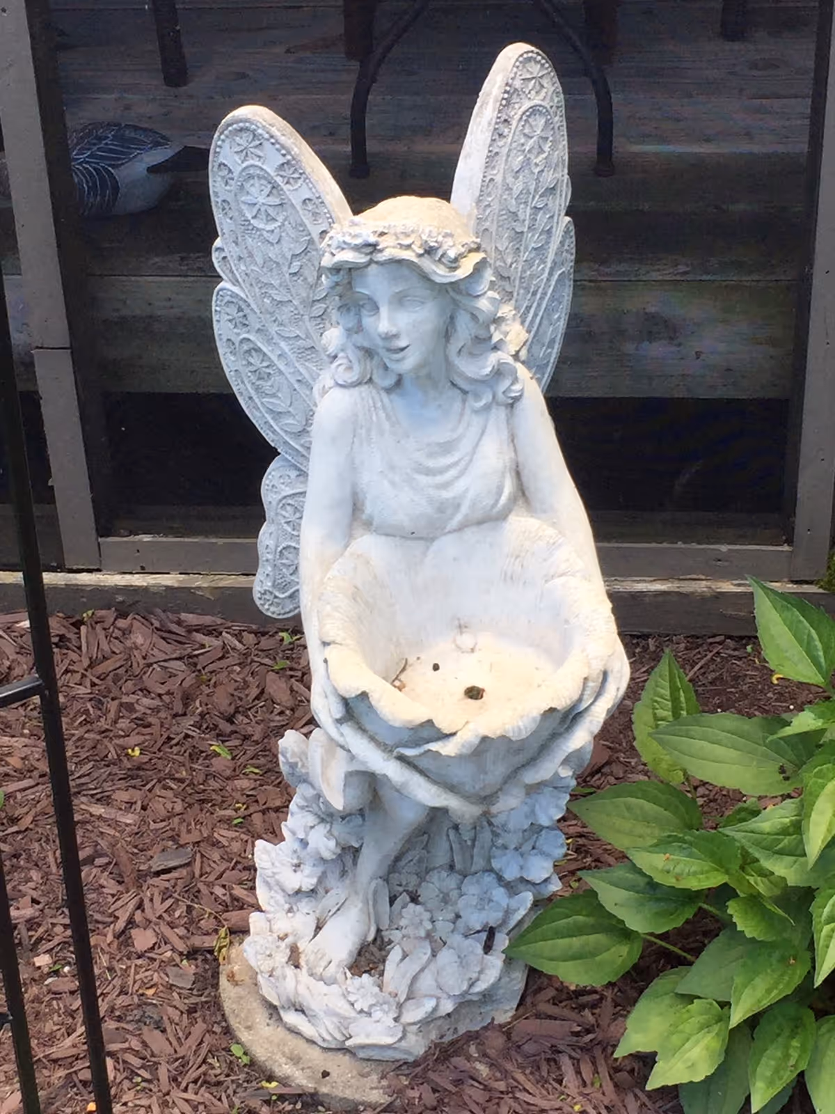 A white stone fairy statue with wings holding a bowl sits in a mulched garden bed next to green plants and a window.