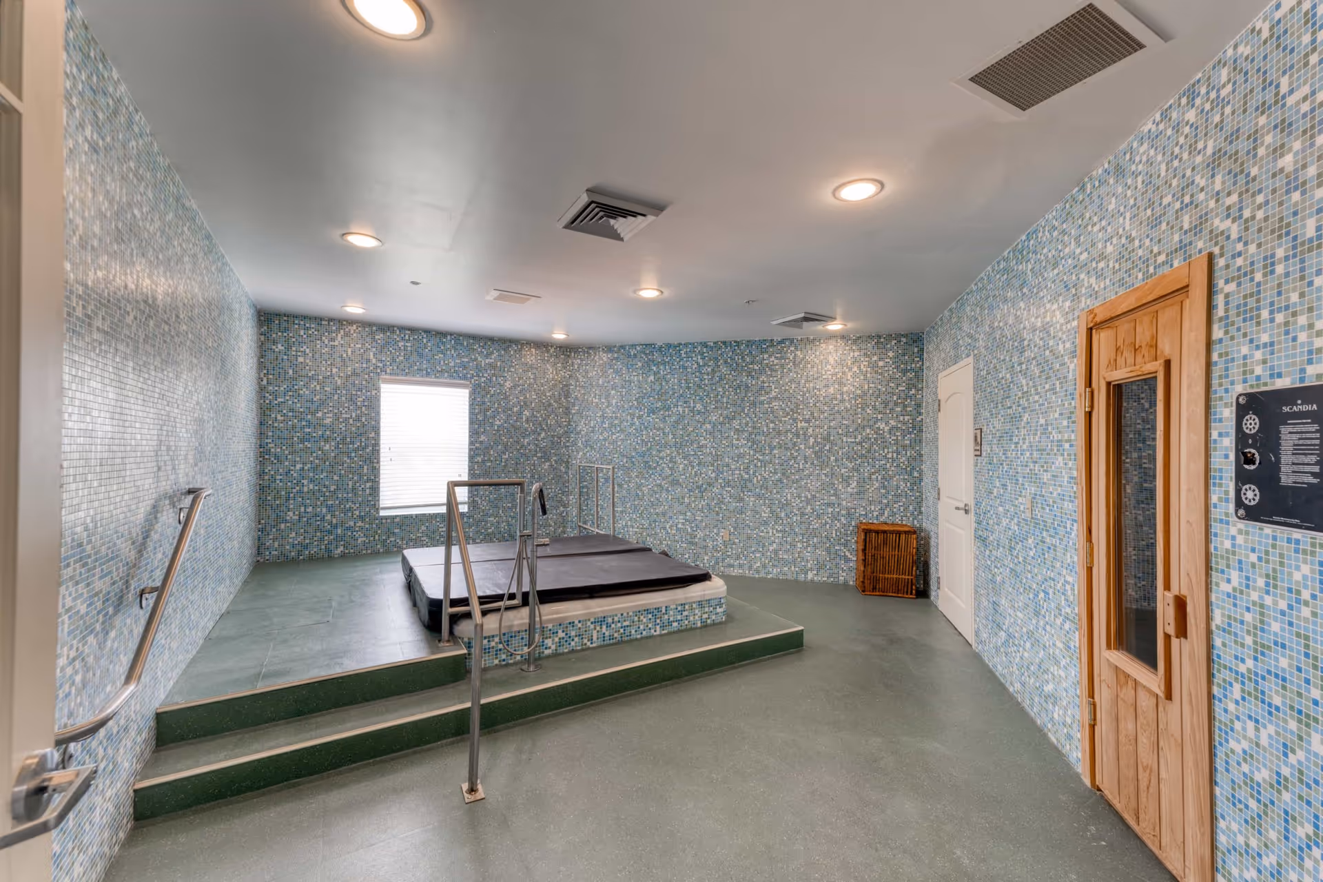 Indoor spa room with blue mosaic tiled walls, a covered hot tub on a raised platform with metal handrails, a wooden sauna door on the right, and a window letting in natural light.