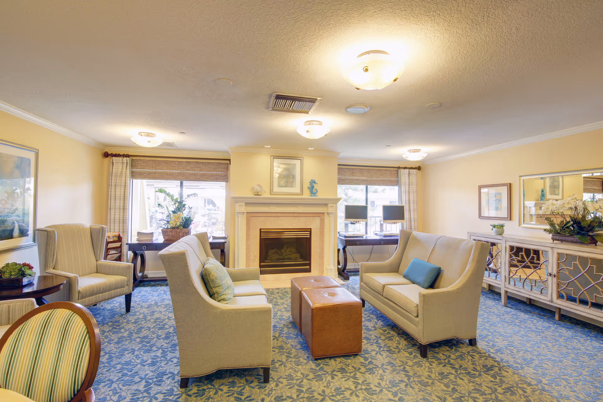 Well-lit senior living lounge with sofas and armchairs arranged around a central ottoman in front of a fireplace.