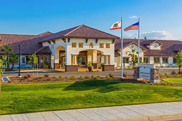 Front exterior of a memory care facility with a covered entrance, two flagpoles, and a manicured lawn.