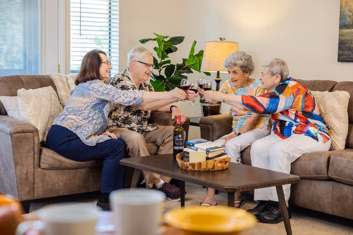 Four elderly people sitting on two brown sofas in a living room, smiling and clinking glasses of red wine together. A coffee table in front of them holds a bottle of wine, books, and a wooden tray. There is a lamp and a green plant in the background.