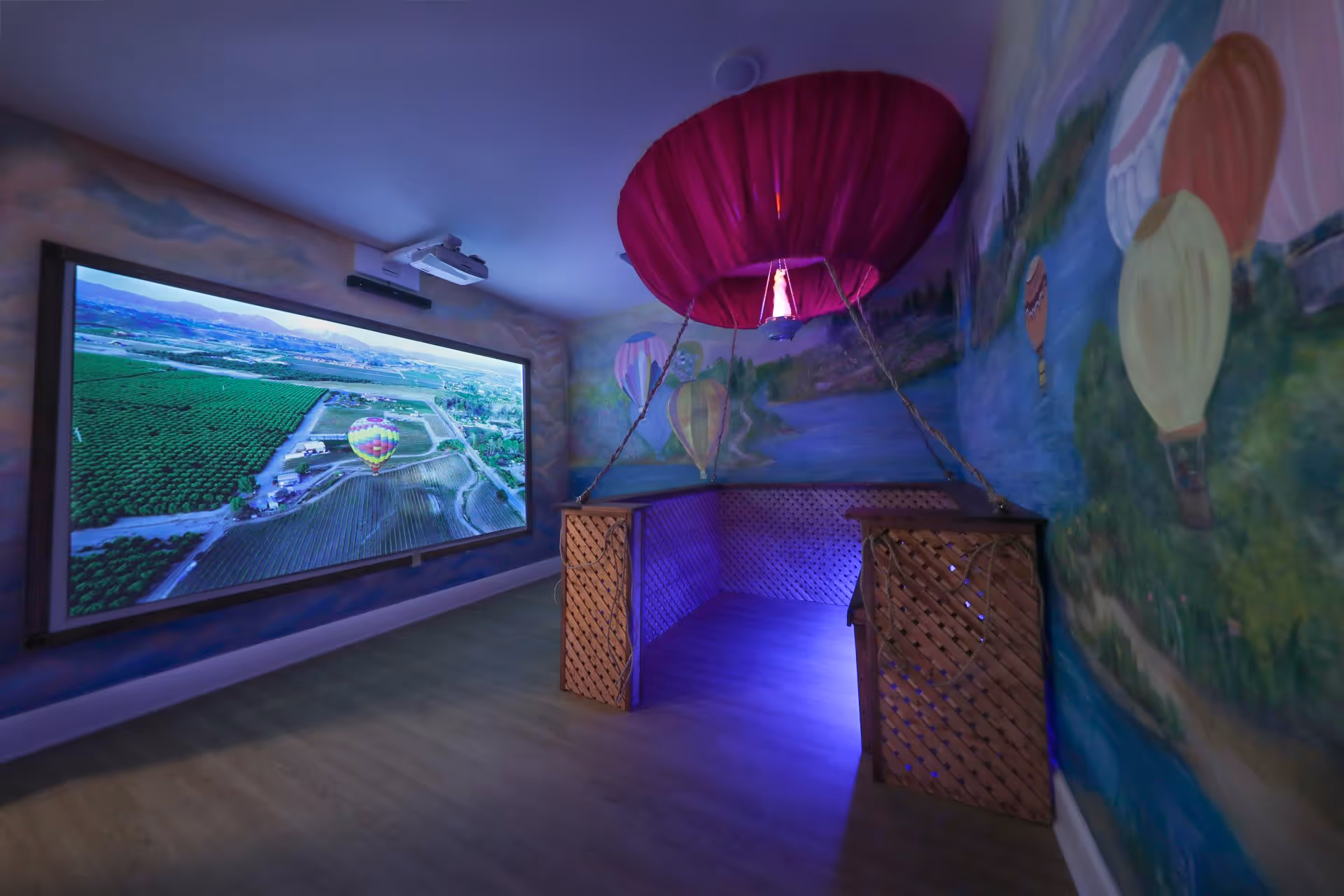 A room with a large screen displaying an aerial view of a landscape with hot air balloons. The room has walls painted with murals of hot air balloons and a decorative hot air balloon basket with a red canopy hanging from the ceiling. The floor is wooden, and the room is softly lit with blue lighting under the basket.