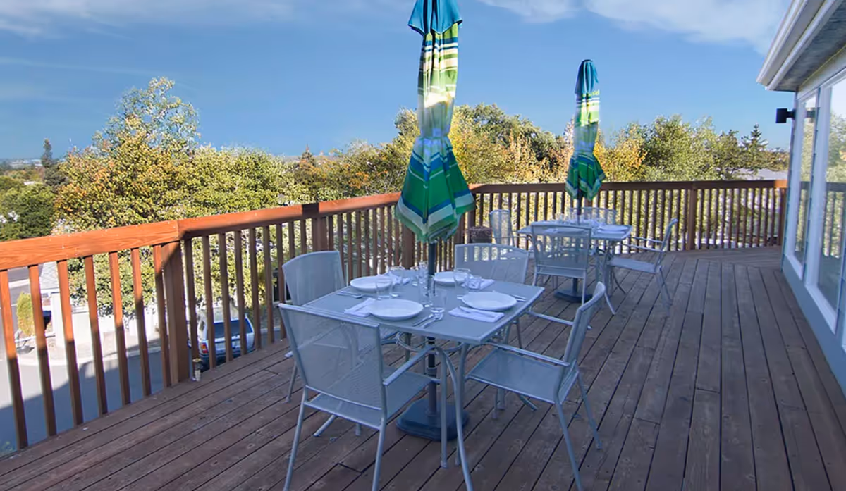 Outdoor wooden deck with two dining tables, each set with white plates, glasses, and napkins. Each table has a closed green and blue striped umbrella. The deck overlooks trees and a parking area under a clear blue sky.