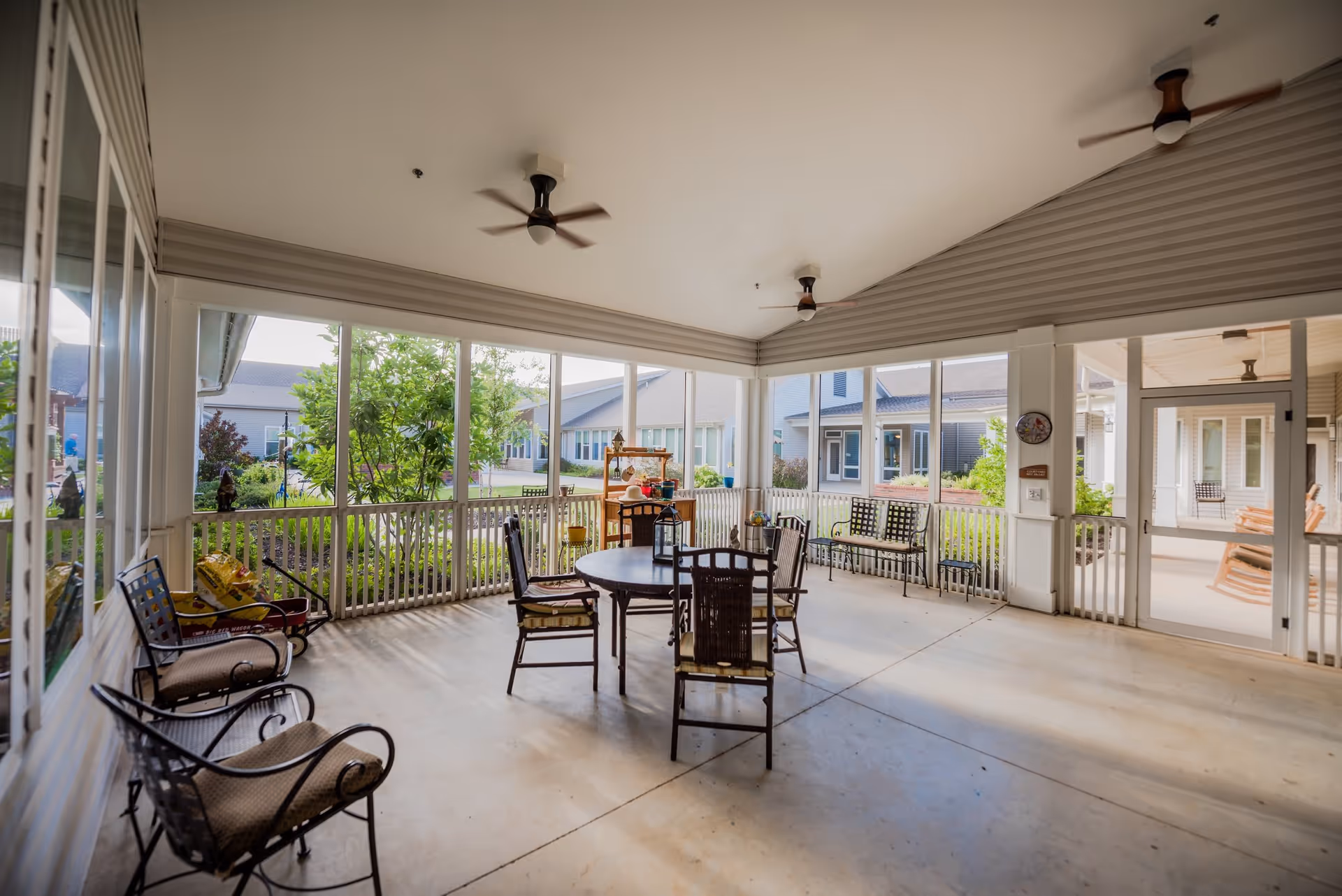 A spacious screened-in porch area with several chairs and a round table in the center. The porch overlooks a garden with greenery and buildings in the background. Ceiling fans are mounted on the white ceiling, and there is a door leading to another outdoor area with rocking chairs.