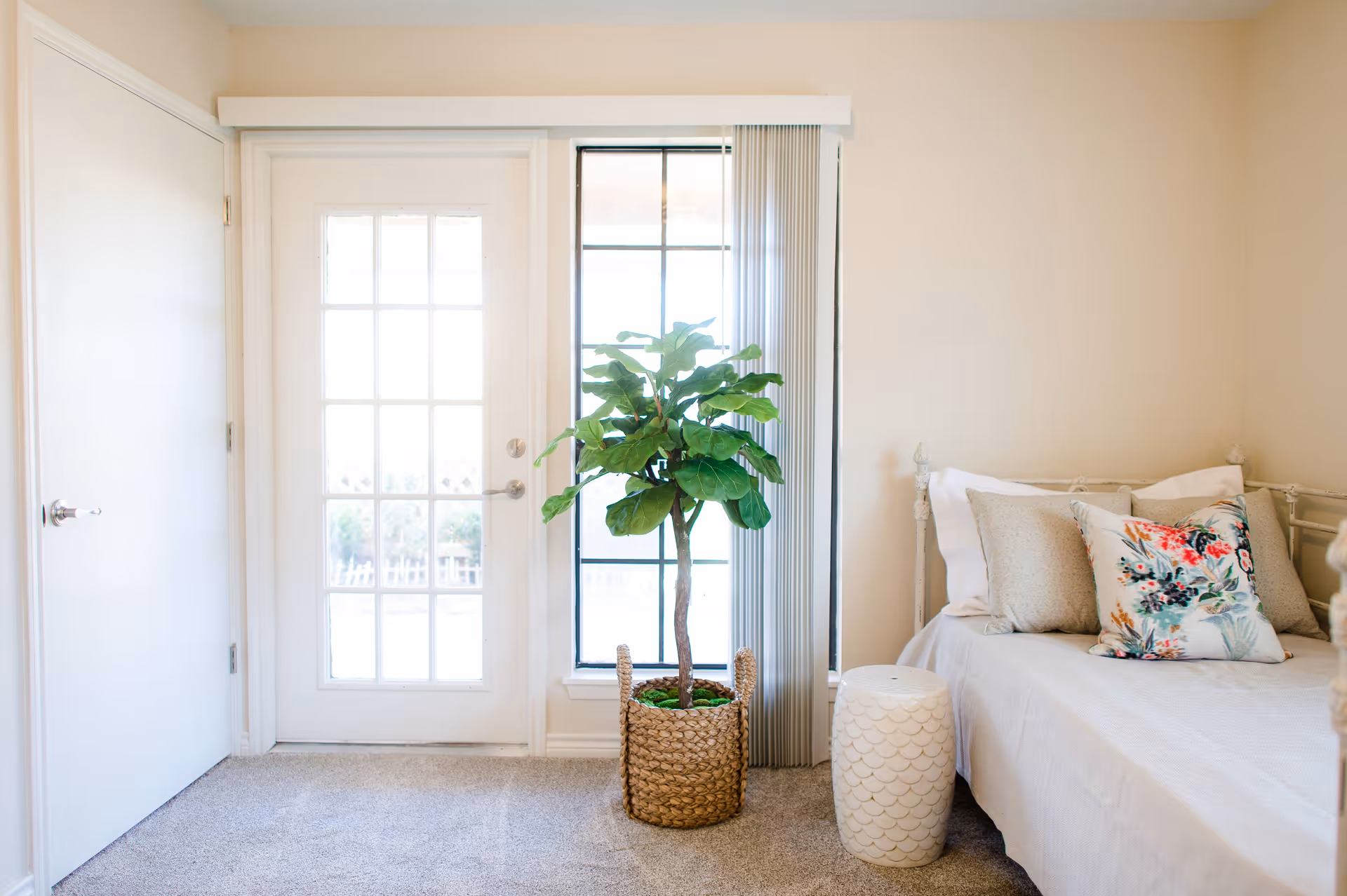 Bright bedroom corner with a potted plant by a glass door and a bed with floral pillows.