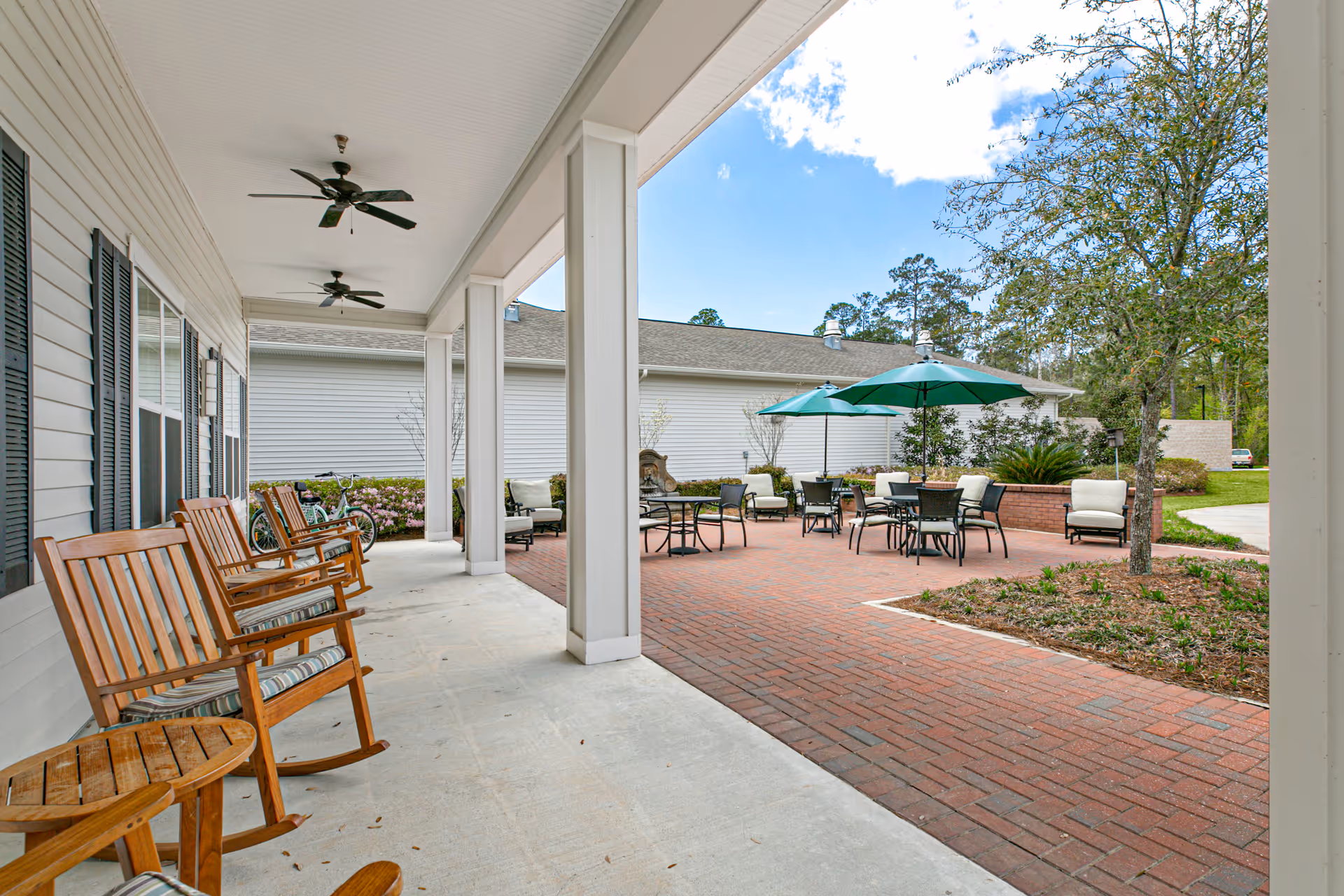 Covered porch with wooden rocking chairs overlooking a brick patio with tables, umbrellas, and outdoor seating.