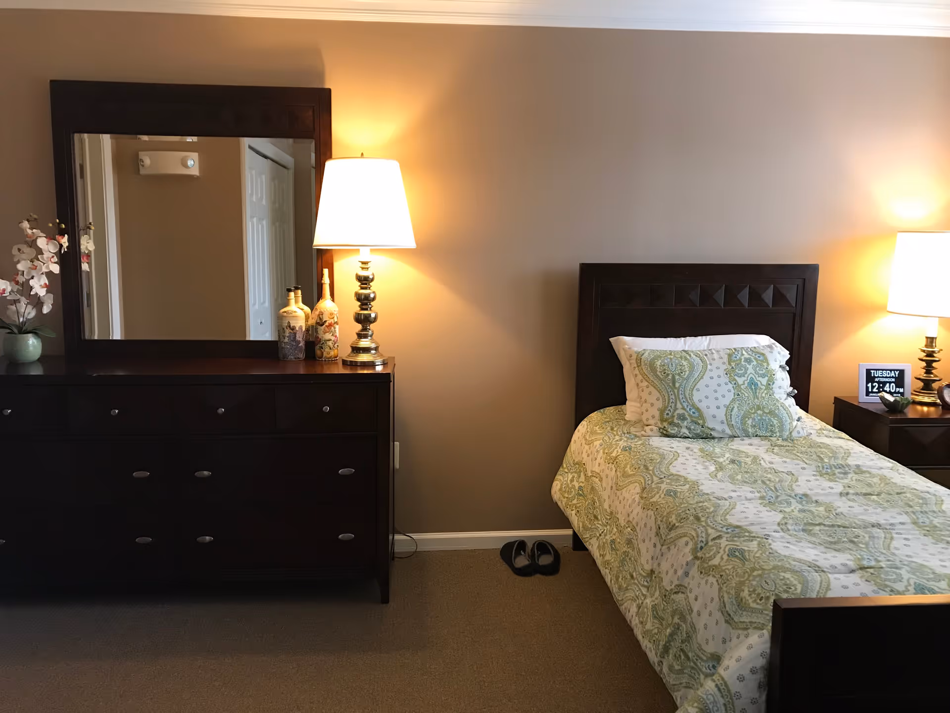 A bedroom with a single bed featuring a green and white patterned bedspread and pillow. There is a dark wooden headboard and a matching nightstand with a lamp and a digital clock showing the time 12:40 PM. To the left, there is a dark wooden dresser with a large mirror above it, decorated with two ornamental bottles and a lamp. A pair of slippers is placed on the carpeted floor next to the bed.