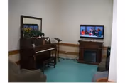 Small communal room with an upright piano, a mirror, seating, and a TV on a stand over a fireplace.