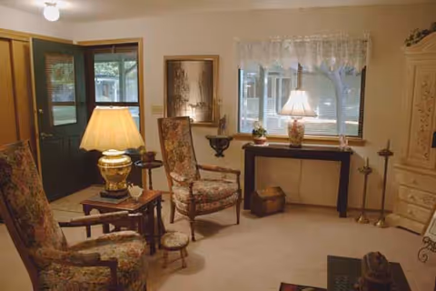 A cozy living room with two floral upholstered armchairs, a small wooden side table with a lamp, a window with lace curtains, a console table with a lamp and decorative items, and a green door partially open to the outside.