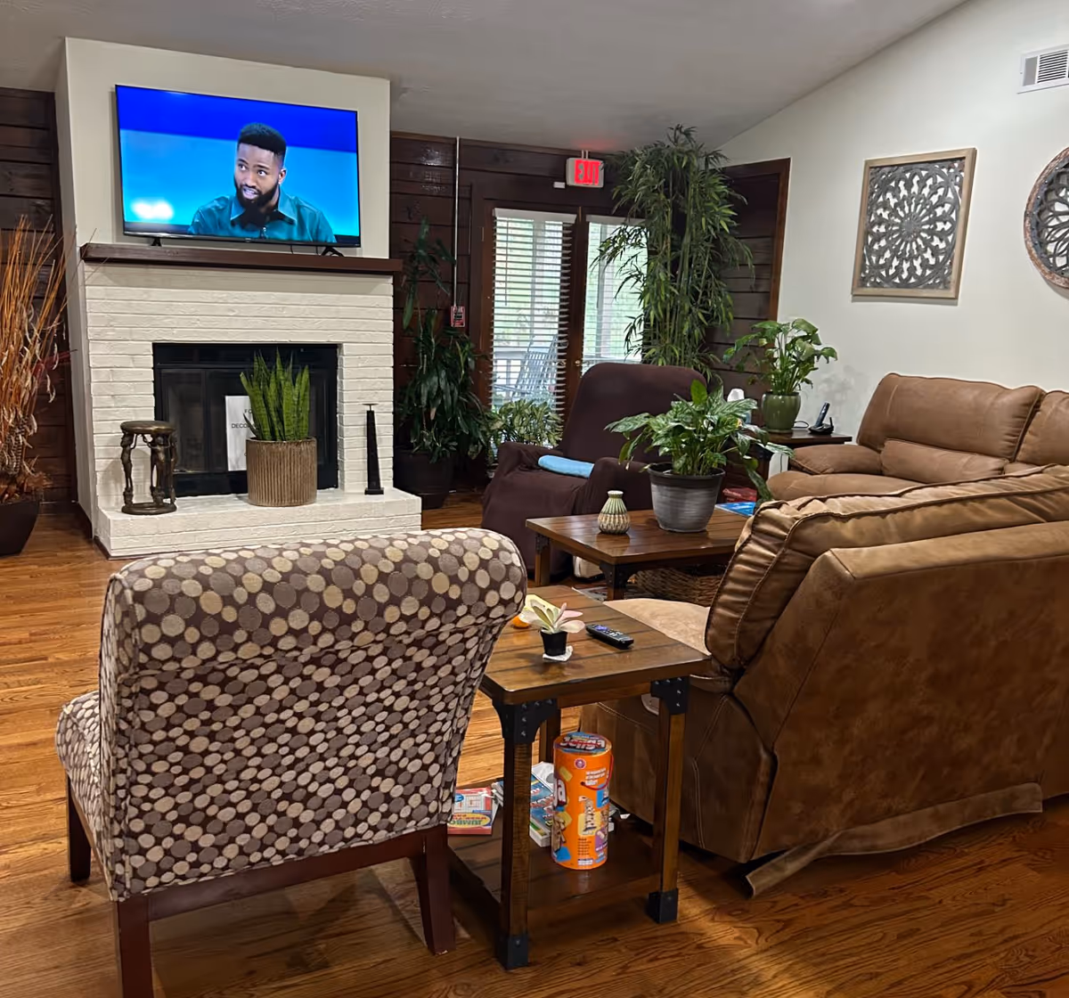 Cozy living room with a patterned armchair, a brown recliner, and a tan sofa arranged around a wooden coffee table and side table. The room features a white brick fireplace with a potted plant on the hearth and a flat-screen TV mounted above it displaying a man speaking. There are several green plants around the room, wooden floors, and decorative wall art. A glass door with blinds and an exit sign is visible in the background.