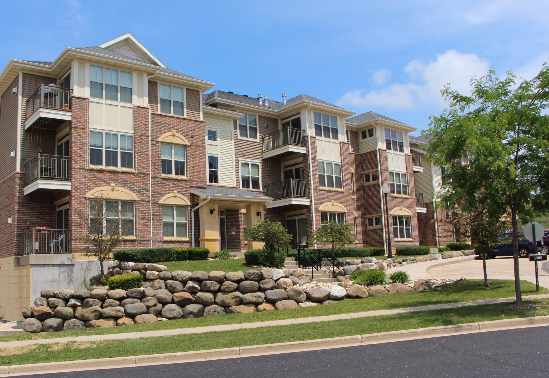 Exterior view of a multi-story assisted living facility building with brick and beige siding, balconies, landscaped rock garden, trees, and a driveway under a partly cloudy sky.