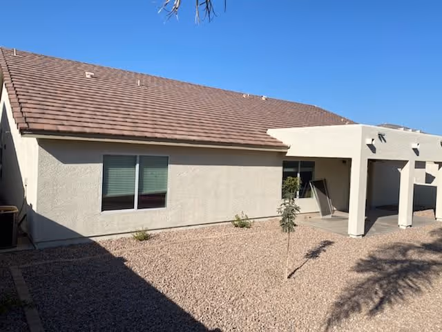 Exterior view of a single-story building with beige walls and a brown tiled roof under a clear blue sky. The ground is covered with small rocks and there are a few small plants and a young tree near the building. There is a covered patio area with an open doorway and a window beside it.