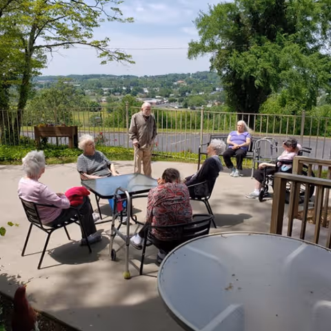 A group of elderly people sitting and standing around outdoor tables on a concrete patio with a scenic view of trees and a distant town under a clear sky.