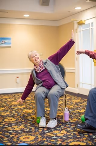 An elderly woman sitting on a chair in a room with patterned carpet, smiling and stretching her arms out to the sides, participating in a seated exercise or activity.
