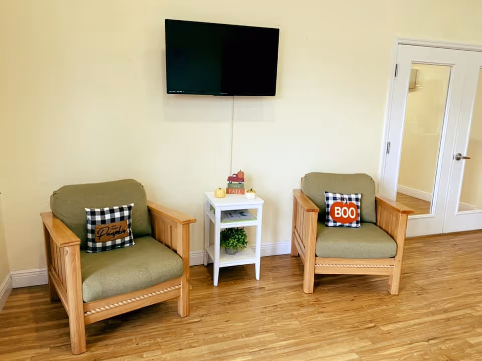 Two wooden armchairs with green cushions and decorative pillows flank a small white side table beneath a wall-mounted TV in a seating area.