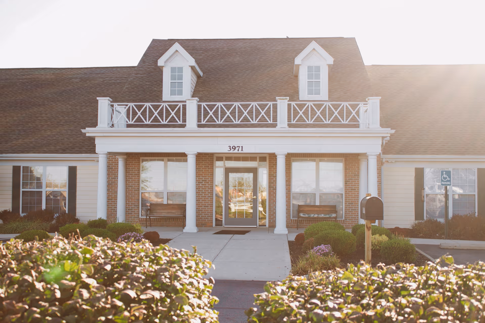 Front entrance of a two-story assisted living building with white columns, a brick facade, address number 3971, benches, and landscaped bushes.