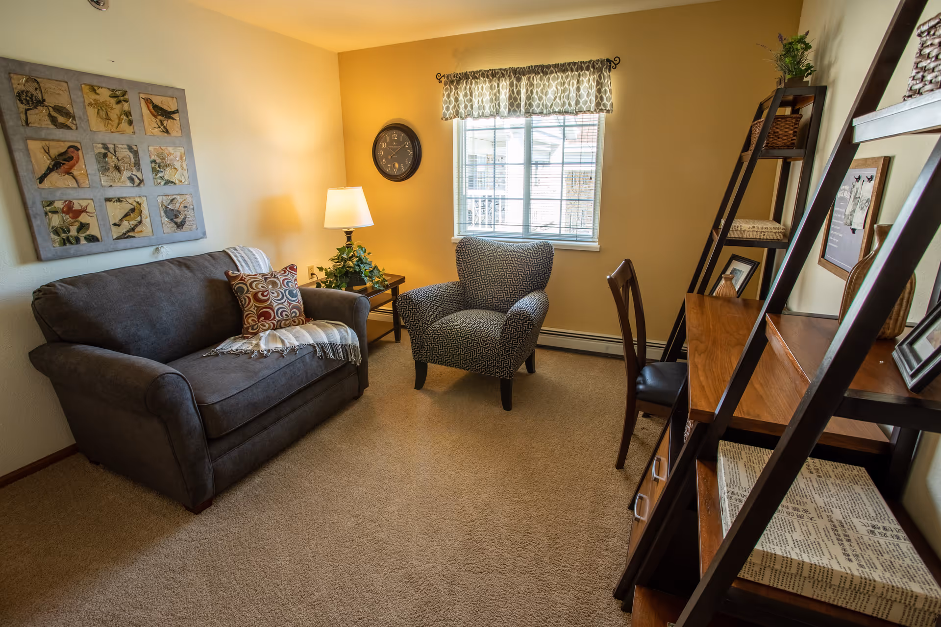 A cozy living room with a gray loveseat adorned with a patterned pillow and throw blanket, a patterned armchair, a wooden side table with a lamp and plant, a wall clock, a window with patterned valance, and a wooden desk with a chair and shelving unit holding decorative items and books.