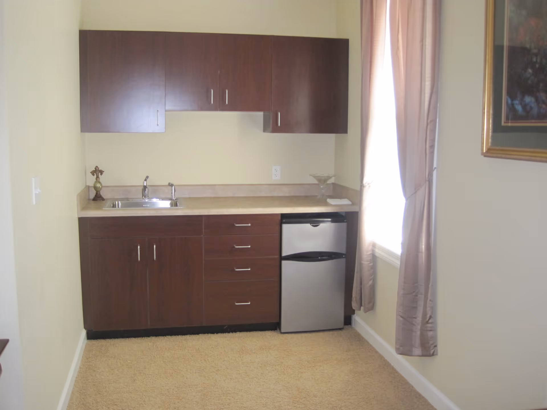 Small kitchenette area with dark wood cabinets, a countertop with a sink, a small stainless steel refrigerator, beige walls, a window with light purple curtains, and a framed picture on the wall.