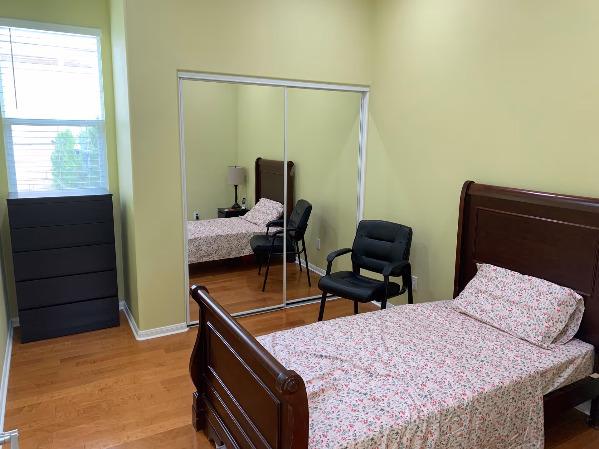 A small bedroom with light yellow walls and wooden flooring. The room contains a single bed with floral bed sheets and a matching pillow, a black cushioned chair, a black chest of drawers, and a nightstand with a lamp. There is a large mirrored sliding closet door reflecting the bed and chair. A window with white blinds is on the left wall.
