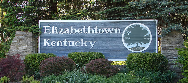 A wooden roadside sign reading "Elizabethtown Kentucky" with a tree emblem set among shrubs and evergreen trees.