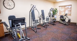 A small fitness room with exercise equipment including a recumbent bike, two weight machines, and a stationary bike. The room has beige walls, a large clock on the wall, a window with blinds, and a potted plant in the corner.