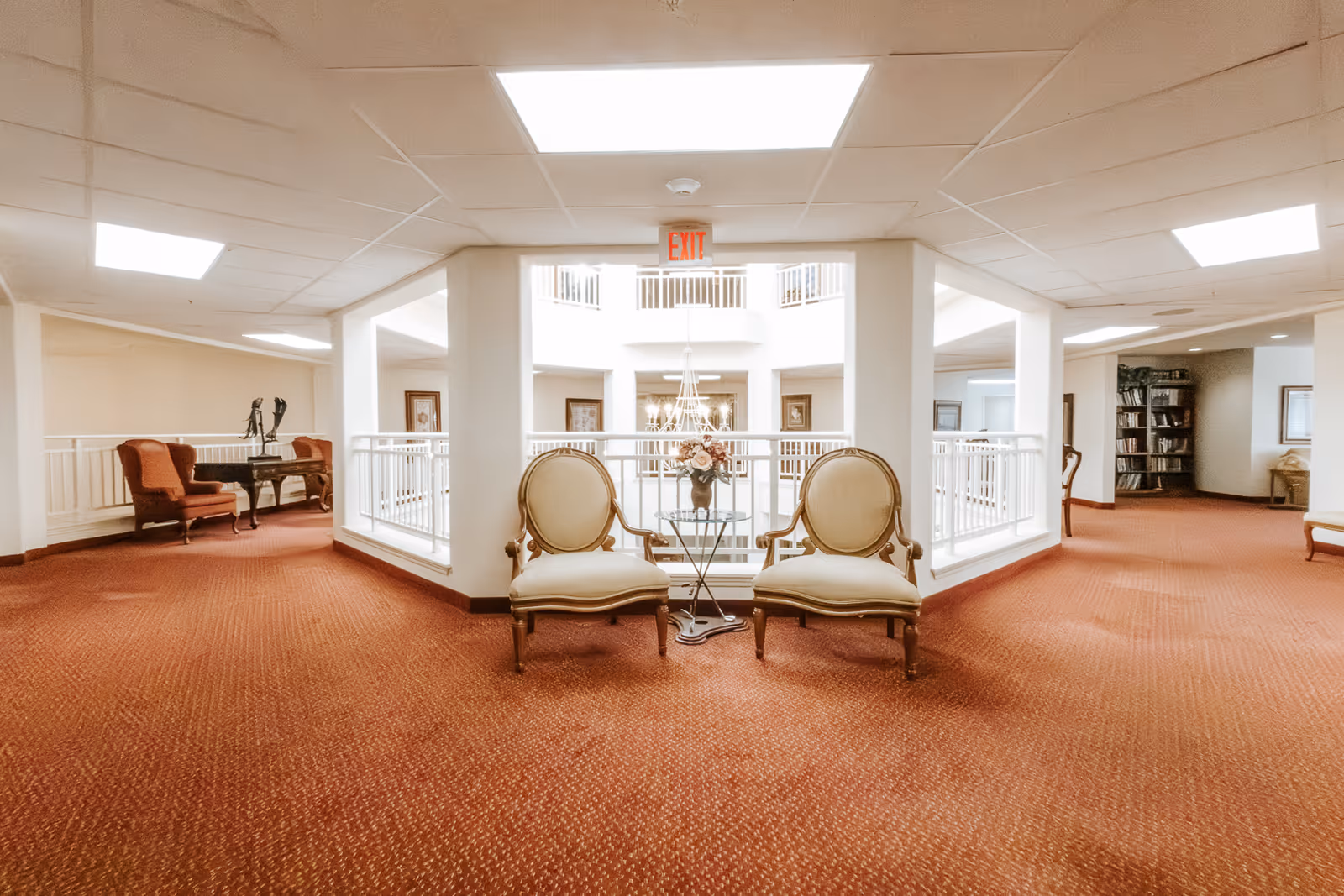 A spacious interior hallway area with red carpet flooring and white walls. Two beige upholstered chairs with wooden frames are positioned around a small glass table with a flower arrangement. The area overlooks a central atrium with a chandelier hanging in the middle. Additional seating and a bookshelf are visible in the background.
