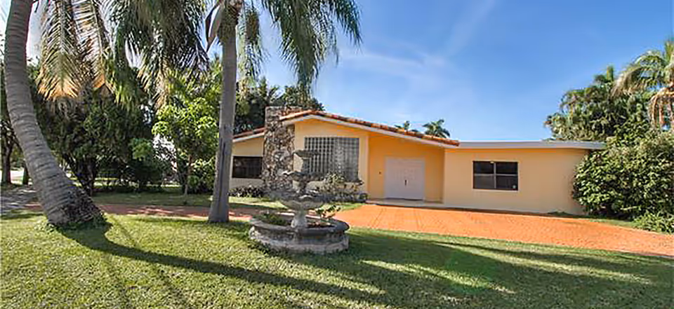 A single-story beige building with a fountain, palm trees, and a green lawn in front.