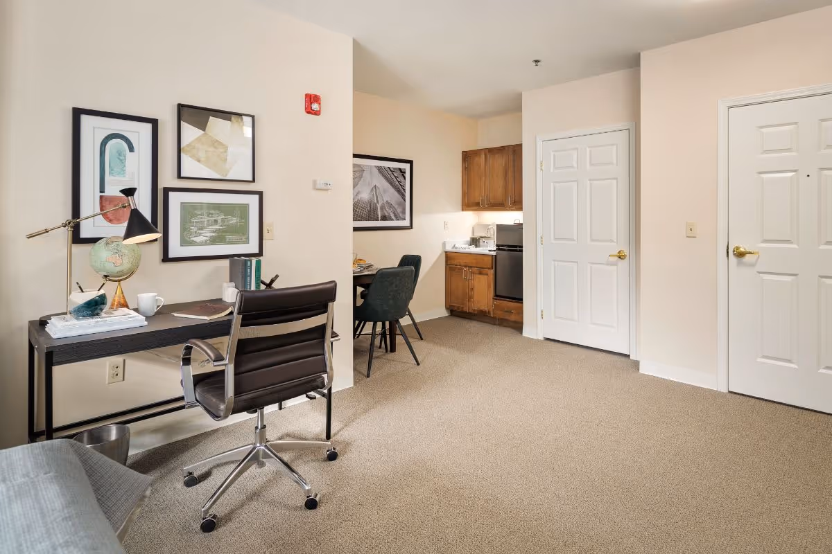 A cozy interior room featuring a small workspace with a black desk, a globe, books, a lamp, and a black office chair on wheels. The wall behind the desk is decorated with framed artwork. In the background, there is a small dining area with a round table and two chairs, and a kitchenette with wooden cabinets, a mini fridge, and a sink. The room has beige walls and carpeted flooring, with two white doors on the right side.