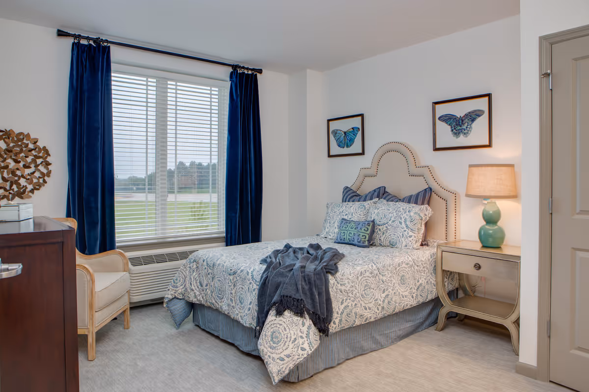 Bright furnished bedroom with a patterned bed and upholstered headboard, nightstand and lamp, armchair, and large window with navy curtains.