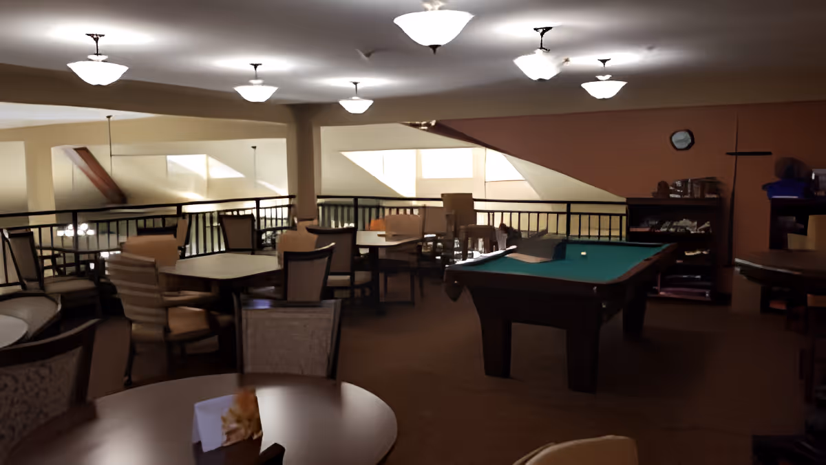 Interior view of a senior living facility common area with multiple tables and chairs arranged for seating. A pool table is prominently placed in the center-right of the room. The space is well-lit with ceiling lights and has a railing overlooking a lower level.