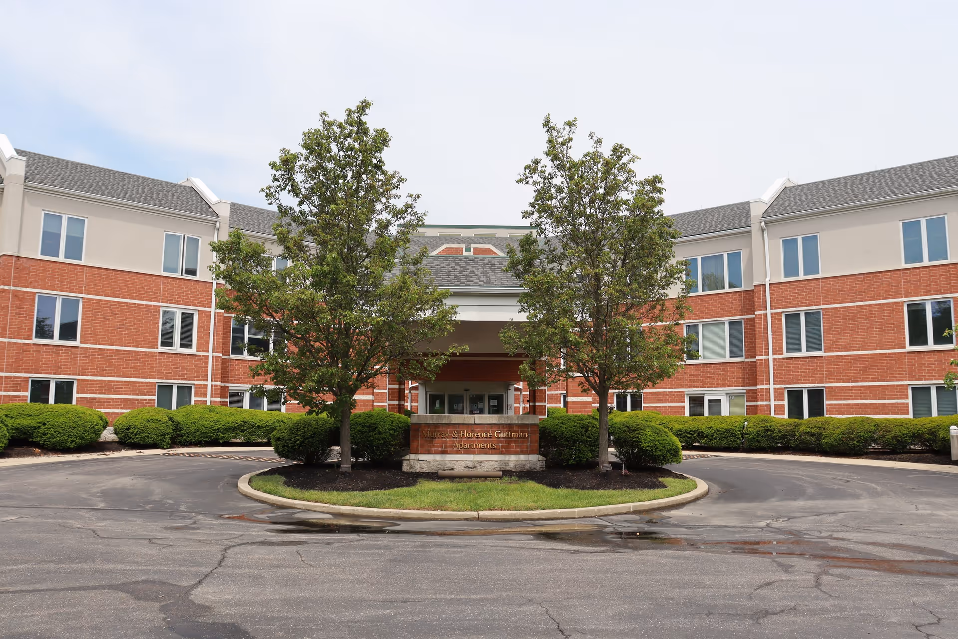 Front exterior view of a three-story brick and beige building with multiple windows, a covered entrance, two trees, and a sign reading 'Murray & Florence Guttman Apartments' in front of the entrance.
