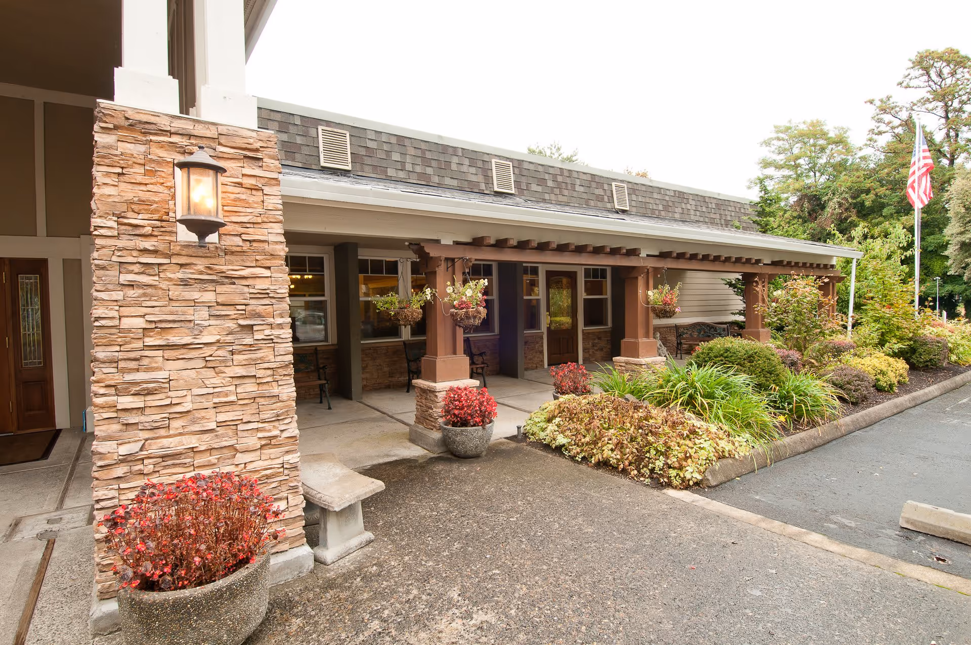 Entrance area of a senior living facility named Royalton Place featuring a covered walkway with stone pillars, hanging flower baskets, potted plants, and landscaped bushes along the driveway. An American flag is visible in the background.