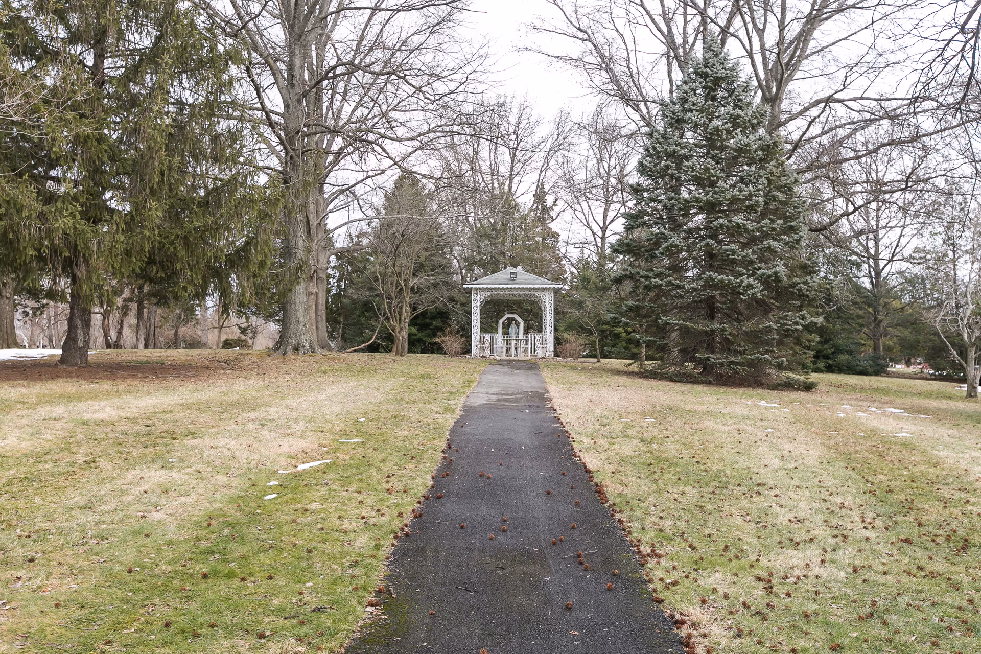 A paved walkway leading to a white gazebo surrounded by trees on grassy grounds.