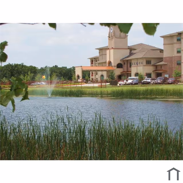 View of a senior living facility building named Amelia Parc with a clock tower, surrounded by greenery and a pond with a water fountain in the foreground.