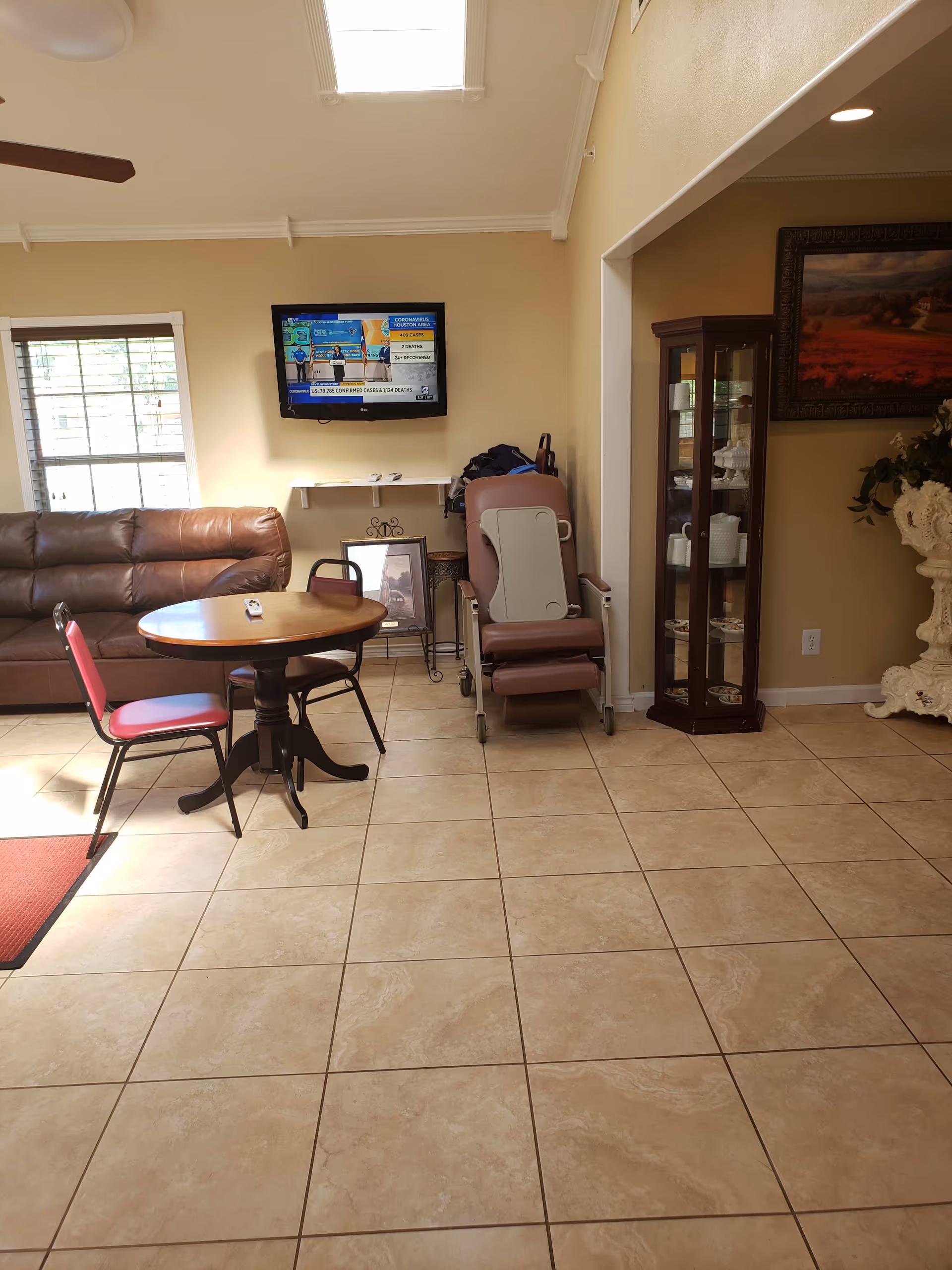 A senior living room with a tiled floor, round table and chairs, leather sofa, wall-mounted TV, and a reclining chair.