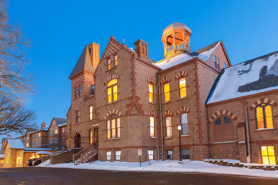 Exterior view of a large historic brick building with multiple windows lit from inside during twilight. The building has a snow-covered roof and surrounding ground, with a clear blue sky in the background. There are steps leading to an entrance and a small parking area with a few cars visible.