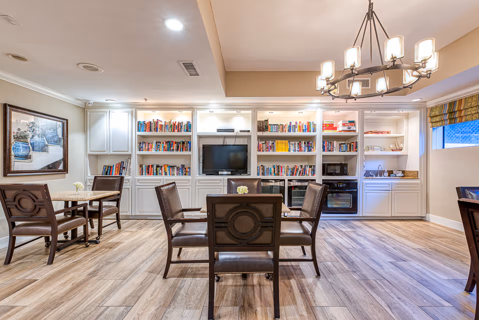 A bright and spacious common area in a senior living facility featuring wooden flooring, multiple tables with chairs, built-in white bookshelves filled with books, a flat-screen TV, and a chandelier hanging from the ceiling. There is a small kitchenette area with a sink, microwave, and oven on the right side, and a framed painting on the left wall.