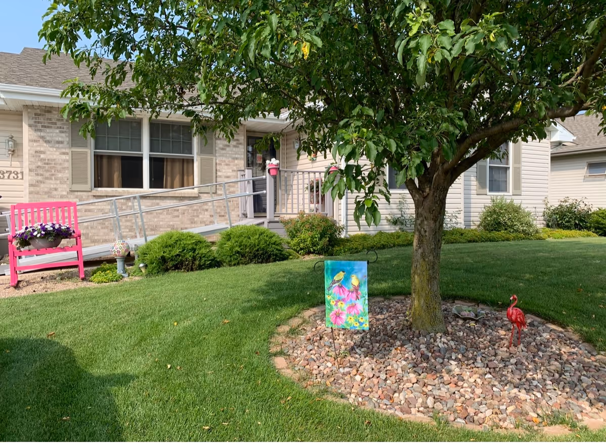 Front yard of a single-story assisted living building showing a tree surrounded by rocks, a colorful garden flag, a pink bench, and an entrance ramp to the door.