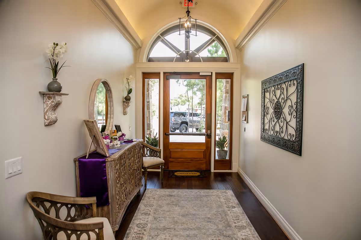 A bright hallway with a wooden front door featuring glass panels and an arched window above. The hallway has light-colored walls, dark wooden flooring, and a patterned rug. On the left side, there is a decorative wooden console table with a purple runner, a round mirror above it, and two small wall shelves holding vases with flowers. A wooden chair with a cushioned seat is also on the left. On the right wall, there is an ornate metal wall decoration. Outside the door, a black vehicle and some greenery are visible.