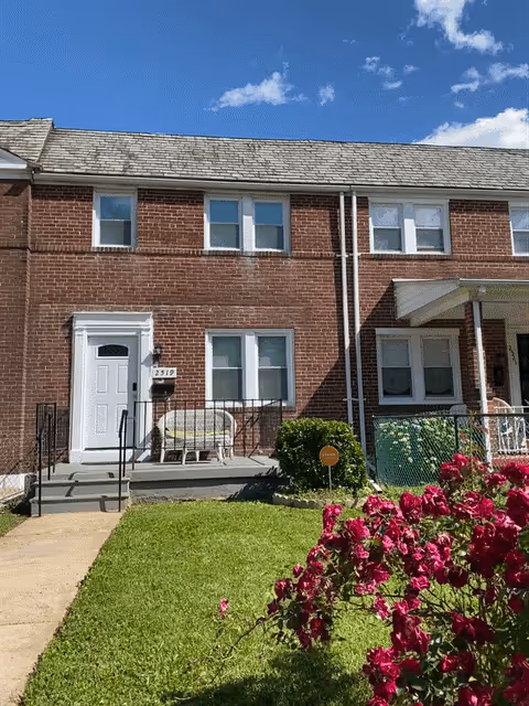 Front exterior view of a two-story brick residential building with a white door, several windows, a small porch with a bench, green lawn, and pink flowers in the foreground under a blue sky with some clouds.