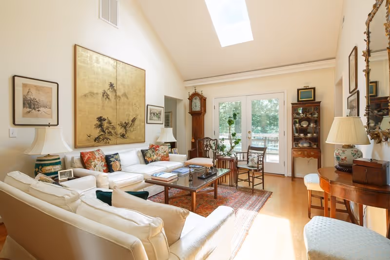 A bright and airy living room with white sofas adorned with colorful cushions, a wooden coffee table with books and decor, a grandfather clock, and glass doors leading to an outdoor area. The room features hardwood floors, a skylight, and various framed artworks on the walls.