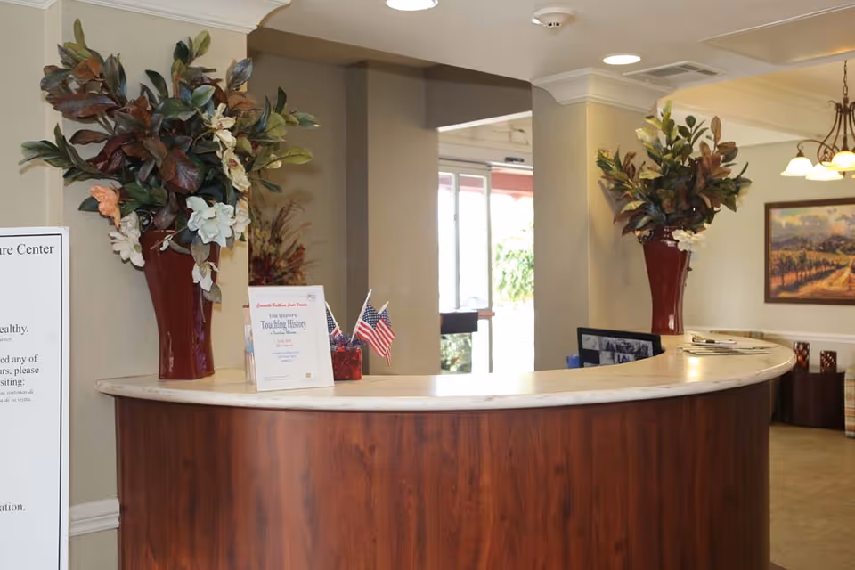 A curved wooden reception desk in a senior living facility lobby decorated with large floral arrangements, small American flags, and informational pamphlets.