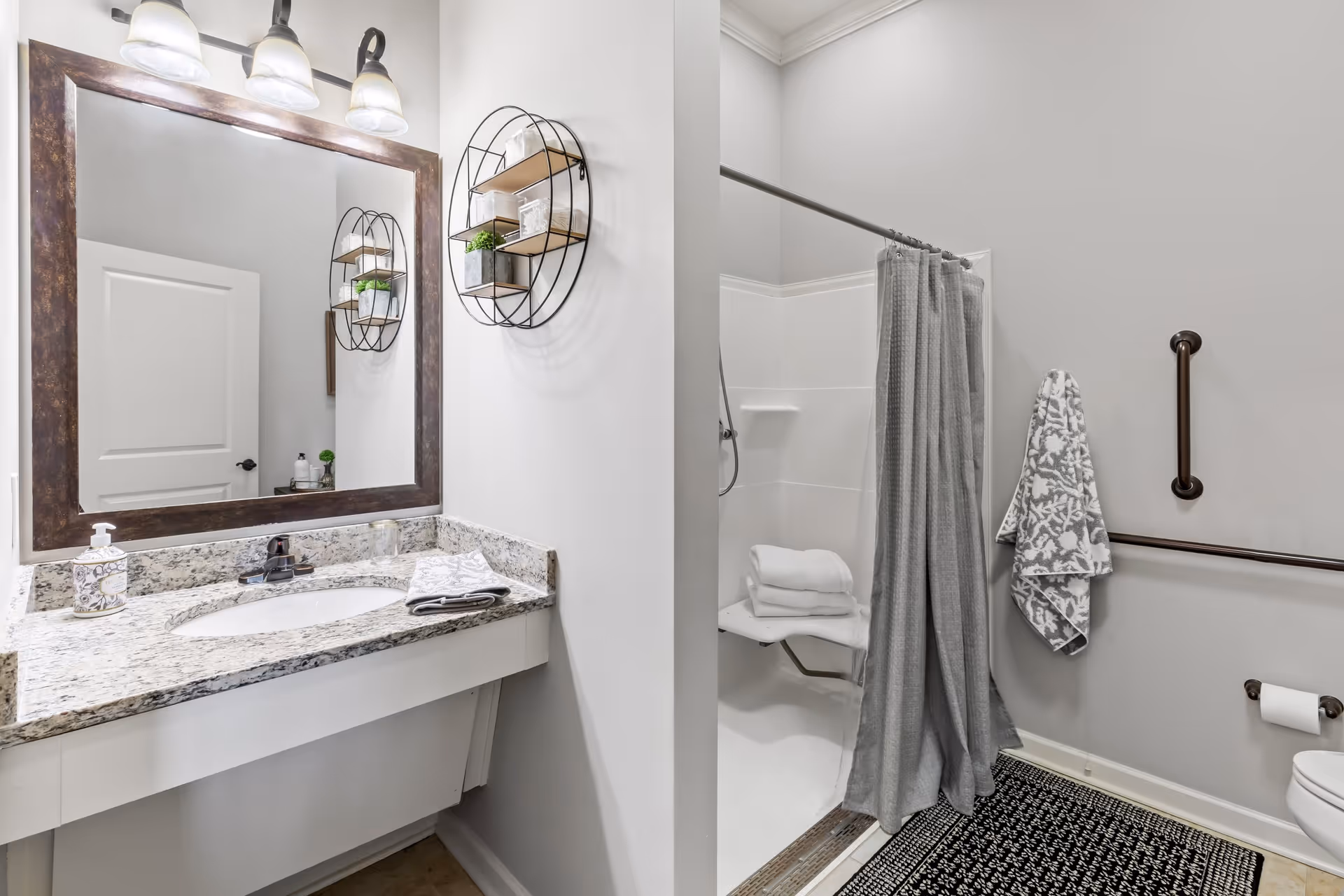 A clean and modern bathroom featuring a granite countertop with a sink, a large framed mirror with three overhead lights, a wall-mounted circular shelf with decorative items, a walk-in shower with a gray curtain, folded towels on a shower seat, grab bars on the walls, a patterned towel hanging, a toilet paper holder, and a black and white patterned rug on the floor.
