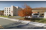 Front exterior of a multi-story senior living building with a covered drop-off entrance, a tree with autumn foliage, and a parked vehicle.